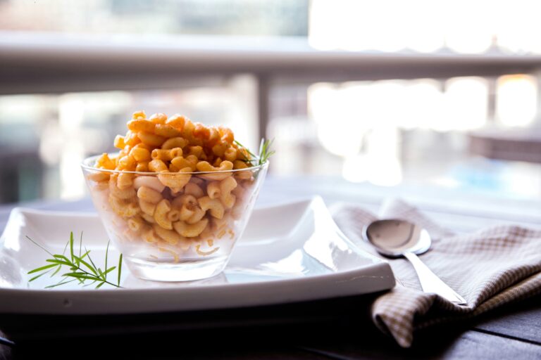 Semangkuk Mac and Cheese creamy dan lezat, disajikan dalam mangkuk kaca transparan dengan piring putih dan hiasan daun rosemary. Tampilan menggugah selera, cocok untuk resep masakan keju yang viral